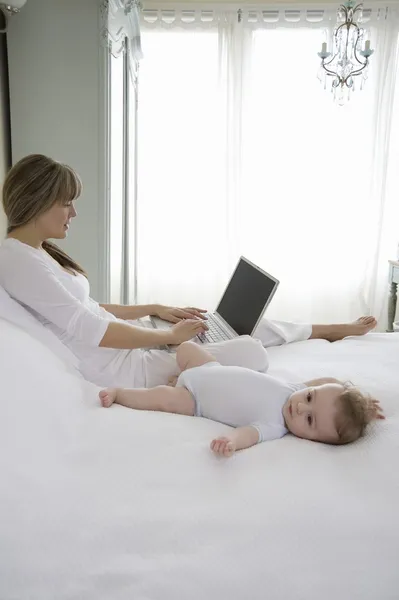 Мать сидит, печатая на ноутбуке с ребенком около нее на кровати — стоковое фото