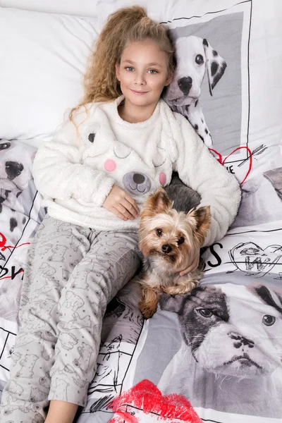 Потрясающие красивая девочка с длинными светлыми волосами лежит на постельного белья в печати собак и лежит рядом с ее собака Йоркширский терьер радостно смотрит в объектив — стоковое фото