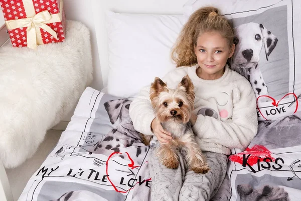 Потрясающие красивая девочка с длинными светлыми волосами лежит на постельного белья в печати собак и лежит рядом с ее собака Йоркширский терьер радостно смотрит в объектив — стоковое фото
