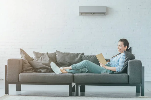 Happy Woman Reading Book Couch Air Conditioner Wall Summer Heat — стоковое фото