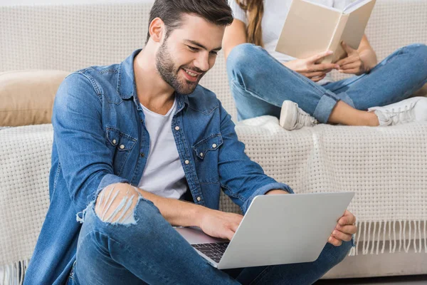 Happy Man Using Laptop Sitting Floor While His Girlfriend Reading — стоковое фото