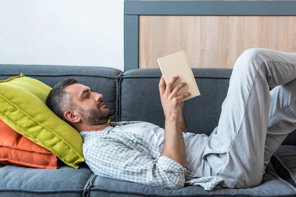 Side View Bearded Man Reading Book While Lying Couch Home — стоковое фото