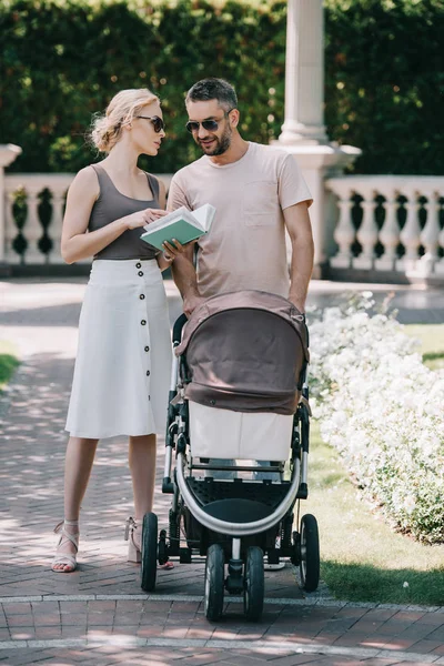 Parents Standing Baby Carriage Park Holding Book — стоковое фото