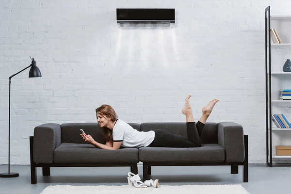 Happy Young Woman Using Smartphone Couch Air Conditioner Hanging Wall — стоковое фото